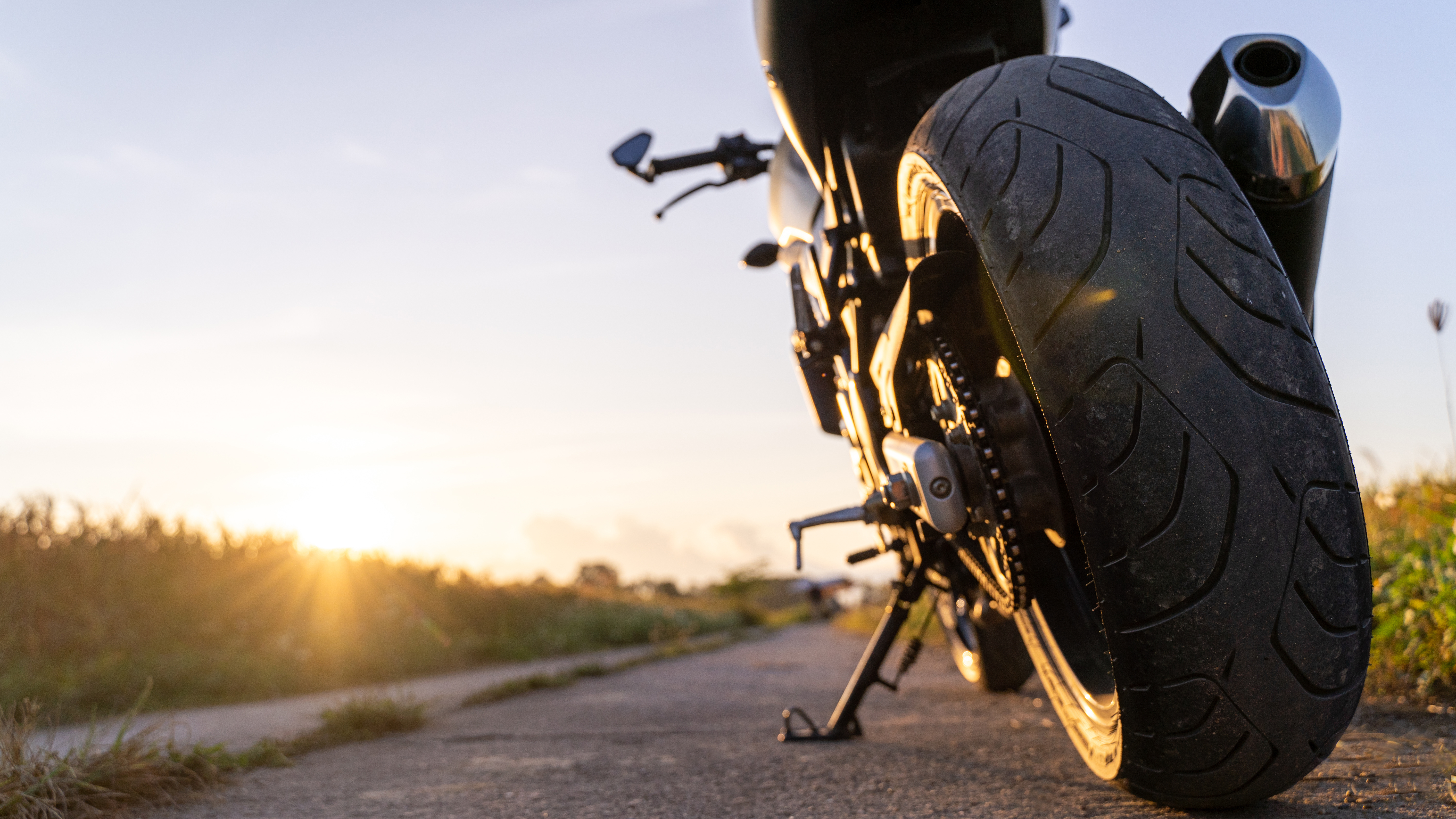 Der Hinterreifen eines Motorrads ist im vorderen Bildbereich zu sehen. Das Krad steht auf dem Ständer auf einer asphaltierten Straße in einer ländlichen Gegend. Im Hintergrund geht die Sonne unter.