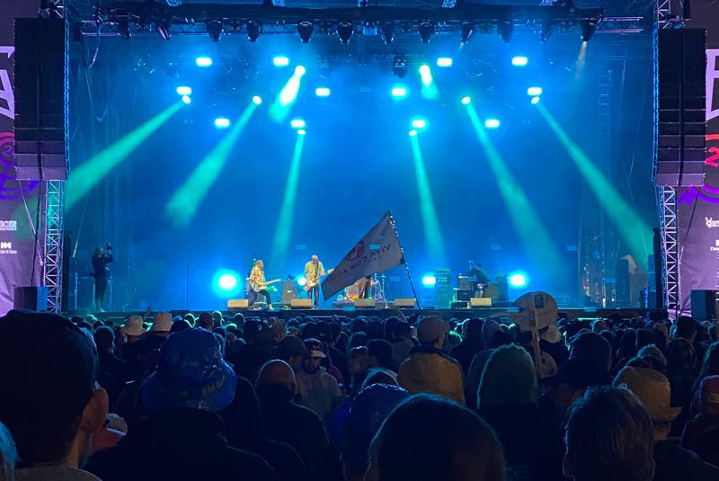 Festival-Stimmung. Im Vordergrund sind Fans vor einer großen Bühne zu sehen. Die Bühne ist mit blauen Schweinwerfern ausgeleuchtet. Auf der Bühne steht eine Band. 