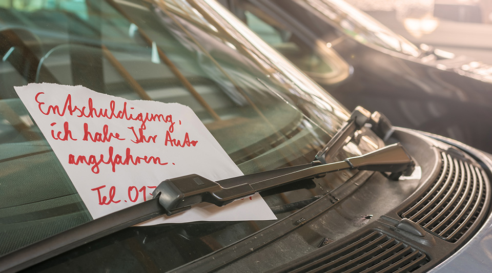 Hinter dem Scheibenwischer an der Windschutzscheibe hängt ein Zettel mit der Aufschrift "Entschuldigung, ich haben Ihr Auto angefahren". Darunter sind Teile einer Telefonnummer zu sehen. 