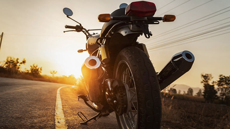 Ein Motorrad ist von hinten zu sehen. Es steht am rechten Straßenrand auf einer Landstraße. Im Hintergrund geht die Sonne unter. 