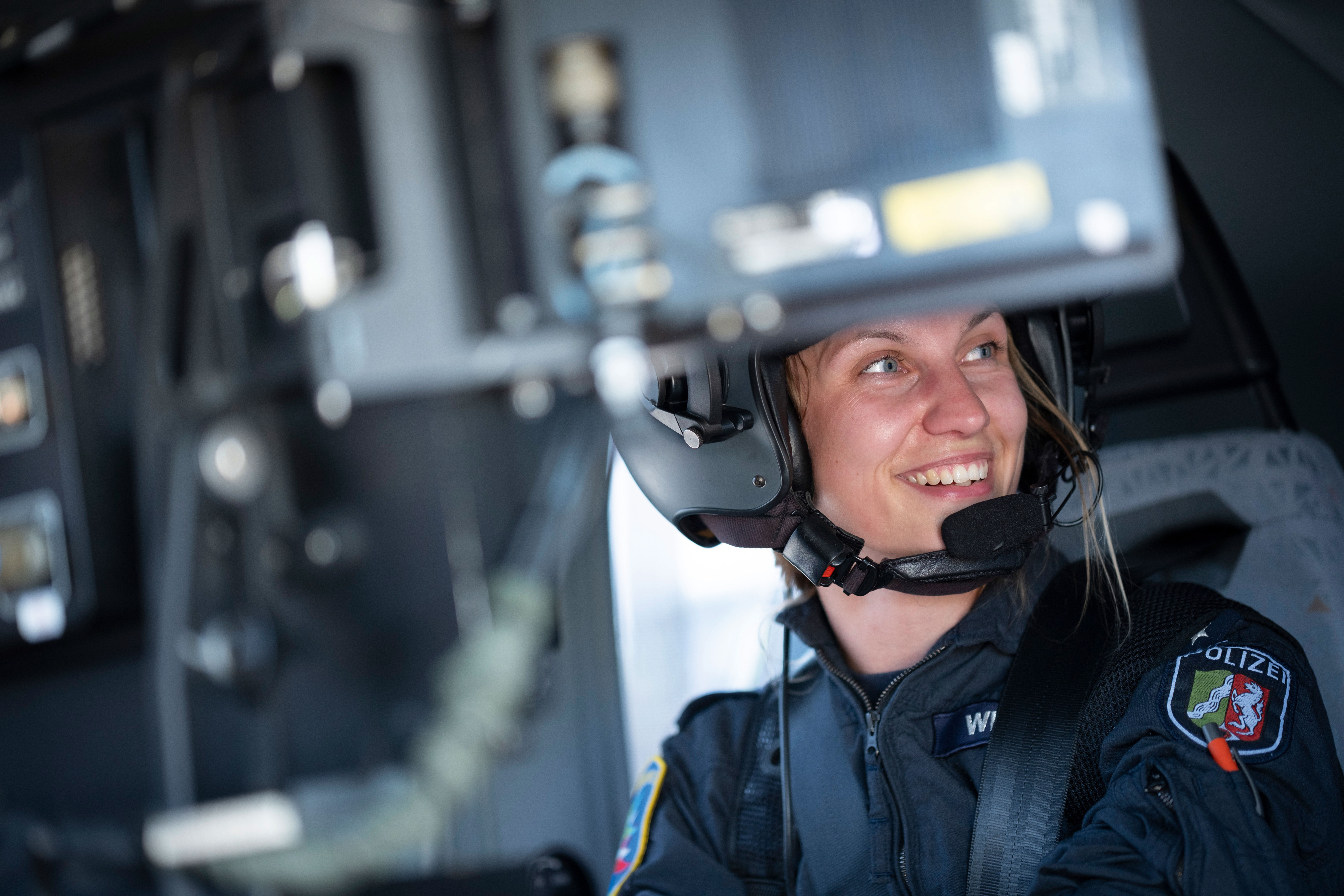 Angelique Wibbeler, Operatorin bei der Polizeifliegerstaffel, sitzt im Cockpit eines Polizeihubschraubers. Sie trägt eine dunkle Uniform mit dem Logo der Polizei NRW auf der Schulter und einen Helm. Sie lächelt.