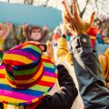 Mehrere Menschen sind von hinten zu sehen, sie tragen bunte Kostüme. Im Hintergrund sieht man verschwommen einen Umzugswagen beim Karnevalsumzug. 