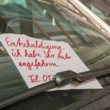 Hinter dem Scheibenwischer an der Windschutzscheibe hängt ein Zettel mit der Aufschrift "Entschuldigung, ich haben Ihr Auto angefahren". Darunter sind Teile einer Telefonnummer zu sehen.