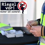 Ein Polizeibeamter, der eine Polizeiweste trägt, steht an einem Stehtisch an einem Infostand der Kreispolizeibehörde Steinfurt. Im Bild ist nur der Oberkörper des Beamten zu sehen. Dieser hält Infomaterial in den Händen. Ein Logo ist oben rechts eingeblendet: Darauf steht Riegel vor! Sicher ist sicherer. 