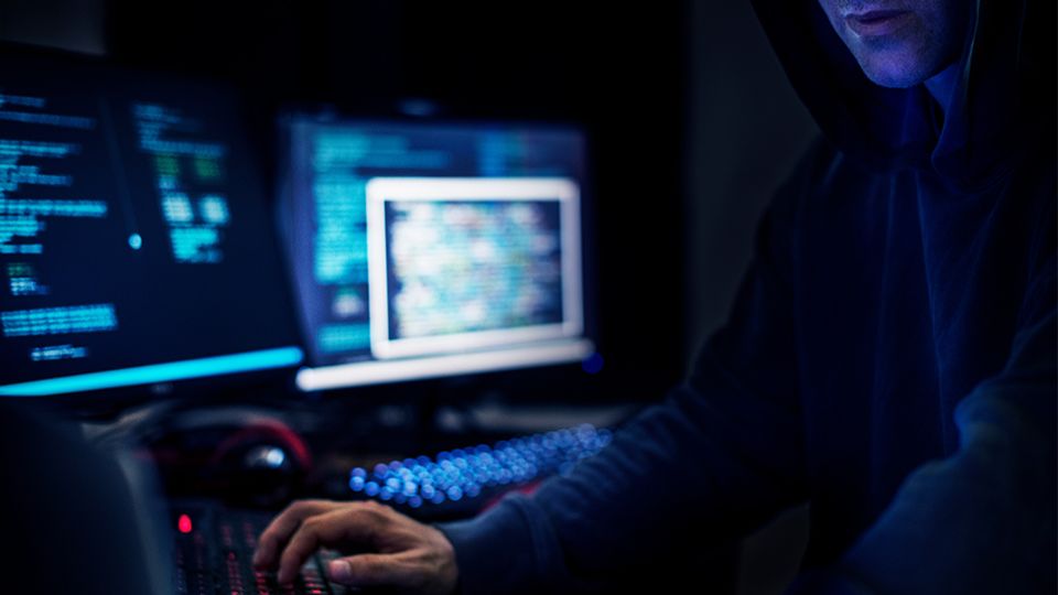 Ein Krimineller, der nur zur unerkannt zu sehen ist, sitzt vor mehreren Computerbildschirmen und tippt in eine Tastatur. 