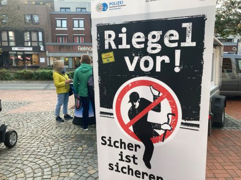 Im Vordergrund steht eine Stellwand mit dem Schriftzug "Riegel vor! Sicher ist sicherer!". Das ist das Plakat der Landeskampagne gegen Einbruchdiebstahl. Im Hintergrund ist der Infostand der Kreisopolizei Steinfurt in Greven zu sehen. 