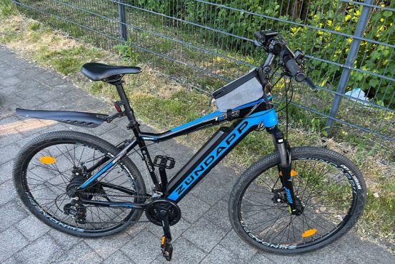 Ein blau-schwarzes E-Mountainbike der Marke Zündapp steht auf einem Weg. Dahinter sind ein Grünstreifen und ein Zaun zu sehen.